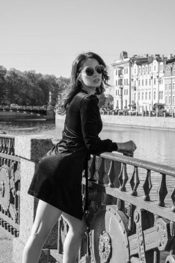 Güzel romantik bir bayanın açık hava portresi. Mükemmel makyaj ve saç stili. Dalgalı saçlı, güneş gözlüklü ve siyah kısa elbiseli bir esmer sahildeki köprüde duruyor..