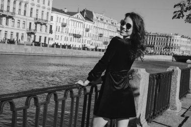 Güzel romantik bir bayanın açık hava portresi. Mükemmel makyaj ve saç stili. Dalgalı saçlı, güneş gözlüklü ve siyah kısa elbiseli bir esmer sahildeki köprüde duruyor..