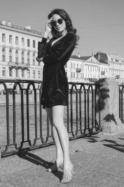 Güzel romantik bir bayanın açık hava portresi. Mükemmel makyaj ve saç stili. Dalgalı saçlı, güneş gözlüklü ve siyah kısa elbiseli bir esmer sahildeki köprüde duruyor..