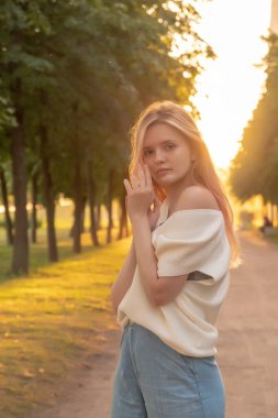 Açık havada, beyaz örgü süveterli güzel sarışın bir kadının, neşeli bir gülümsemeyle kameraya bakarken, gün doğumunda arka planda...