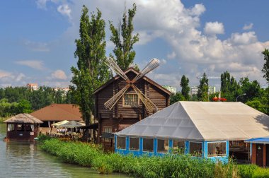 Krasnodar, Rusya - 01 Haziran 2015: Nehrin kıyısında bir yel değirmeni olan ev, diğer binalar ve ağaçlarla çevrili mavi bulutlu gökyüzüne karşı