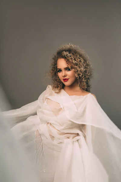 Elegant curly woman in white chiffon dress on grey background