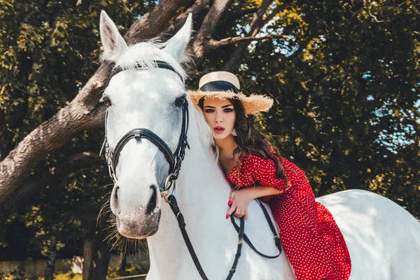 Young stylish woman in red dress posing on white horse