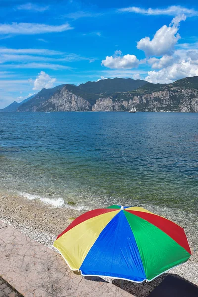 Ombrellone colorato sulla riva del lago di garda, barca a vela sul lago e — Foto Stock