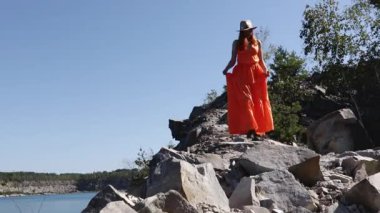 Turuncu elbiseli güzel bir kız göl kenarındaki kayalık bir uçurumda yürüyor. Vahşi doğada kadın güzelliği kavramı. Rahatlama ve huzur yeri