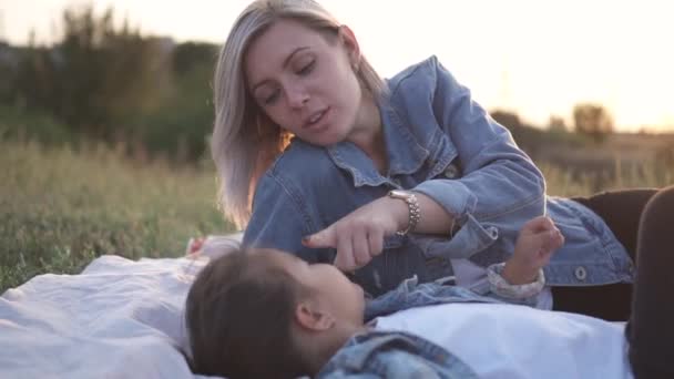 混血家庭 欧洲母亲和亚洲女儿在日落时在公园里度过的时光 他们躺在地上微笑着亲吻对方 温暖的春天或夏季 — 图库视频影像