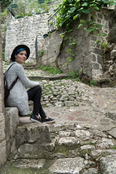 Una chica con un sombrero negro se sienta en escalones de piedra cerca de las carreteras pavimentadas en la vieja ciudad europea . — Foto de Stock
