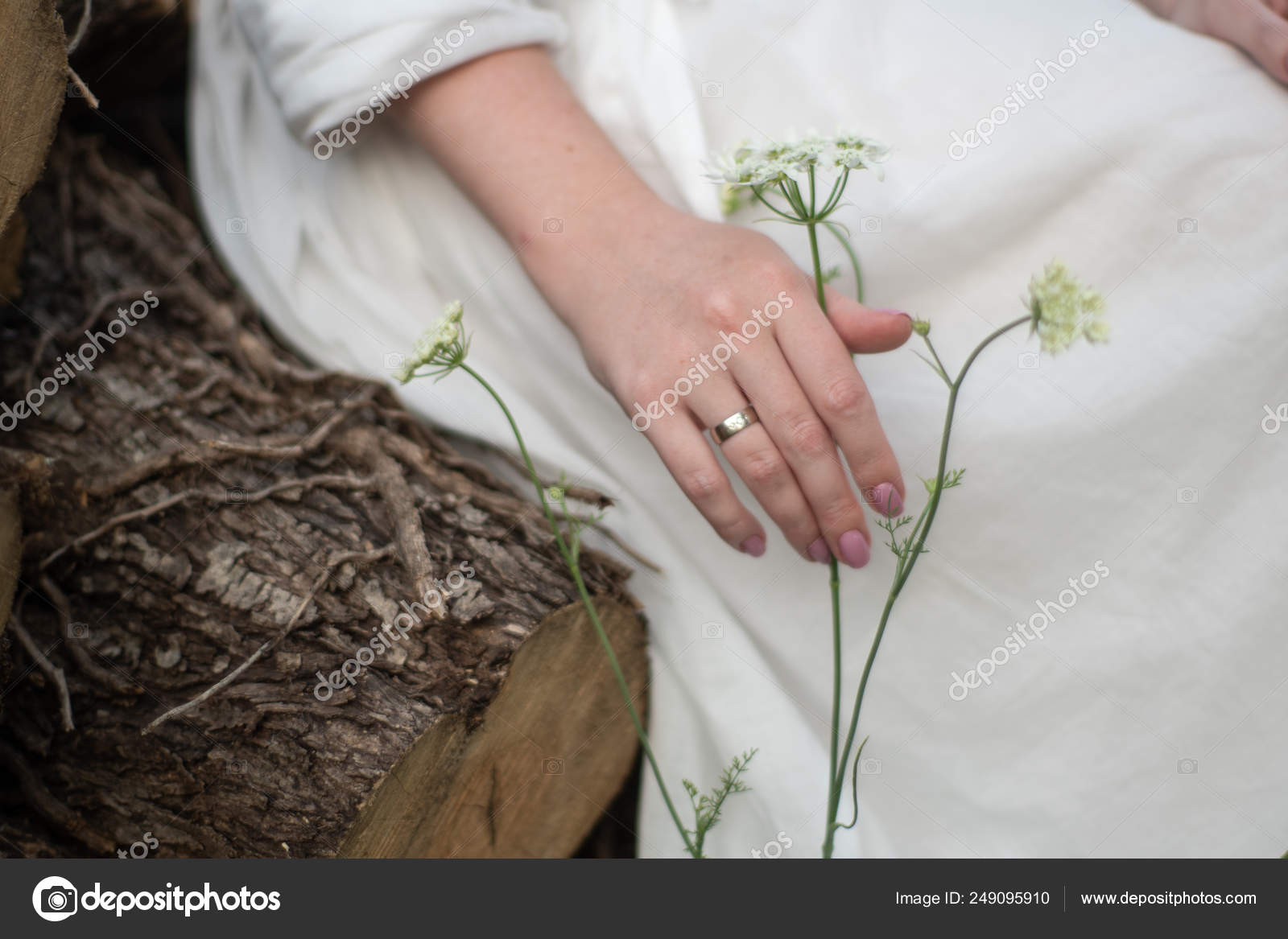 A Girl In A White Dress Sits On A Wooden Log And Touches A Flower