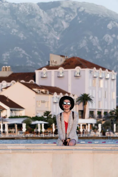 Una chica con gafas de sol y un sombrero negro está de pie en un muelle cerca del mar en una ciudad turística europea en el fondo de casas de lujo y palmeras. Viajes y vacaciones — Foto de Stock