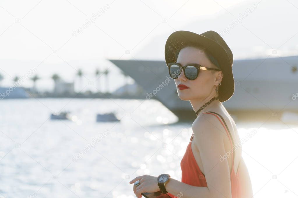 The girl in sunglasses and a black hat on the background of yachts in the resort European city looks into the distance. Travel and Vacation