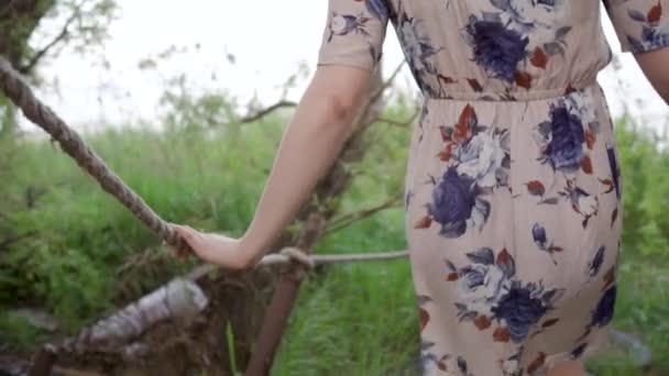 Une jeune fille aux cheveux courts portant une robe avec des fleurs passe pieds nus sur le sol et le pneu traverse les saules. Tournage depuis le dos