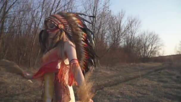 先住民族のインディアンの頭飾りと衣装を着た美しい少女の日没の儀式的なダンス 川の近くの木の中で彼女の手の中にインドの草とカラフルな化粧で ソルガストラム ナッツ — ストック動画