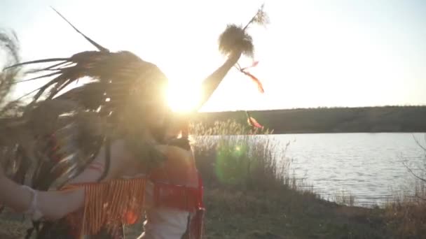 ネイティブアメリカインディアンの頭飾りとカラフルな化粧をした衣装を着た美しい女の子は 日没時にインド草で体に沿って儀式的な動きを実行します ソルガストラム ナッツ — ストック動画