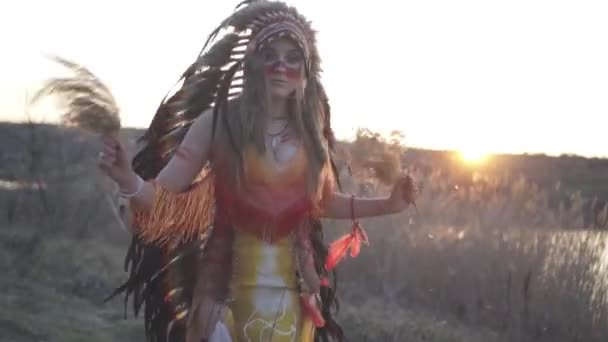 美丽的女孩穿着土生土长的印第安人头饰和服装 五颜六色的妆容 手握印第安草 在夕阳西下表演着仪式舞 山核桃属 — 图库视频影像
