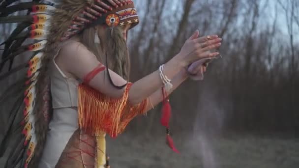 美丽的女孩穿着土生土长的印第安人头饰和服装 在日落时的仪式性舞蹈中妆容五彩斑斓 她把袋子和粉红的粉末从里面挤出来 — 图库视频影像