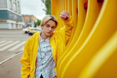 Sarı rüzgarlık ve beyaz gömlekli bir adam sokakta sarı bir yapının yanında duruyor..