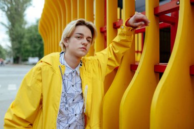 Sarı rüzgarlık ve beyaz gömlekli bir adam sokakta sarı bir yapının yanında duruyor..