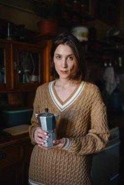 Bej örgü kazaklı üzgün bir kız elinde kahve makinesiyle eski bir ahşap büfenin yanında duruyor.