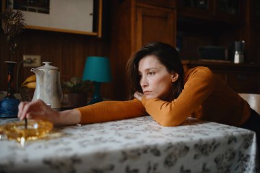 Kahverengi kazaklı üzgün bir kız mutfakta oturuyor ve kafası masanın üzerinde dinleniyor.