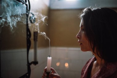 Banyodaki kız, aynanın önünde sönmüş dumanı tüten bir mum tutuyor.