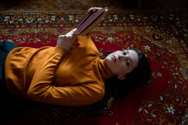 Turuncu kazaklı üzgün bir kız odada kırmızı bir halıya uzanıyor ve elinde bir kitap tutuyor.