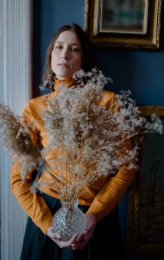 Turuncu kazaklı bir kız mavi duvarlı bir odada duruyor ve önünde kurumuş kır çiçekleriyle vazo tutuyor.