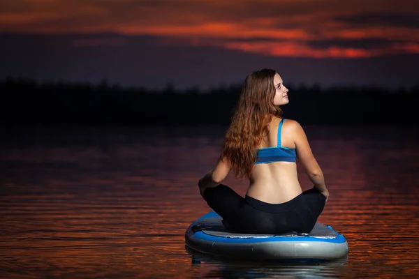 stock image Beautiful slim fit girl with long brown hair exercising yoga on paddleboard in the dark, colorful sunset on scenic lake