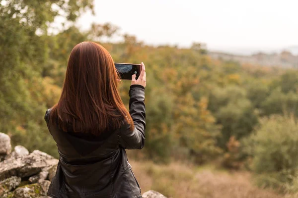 Una joven pelirroja toma fotos con su teléfono móvil en un bosque — Foto de Stock