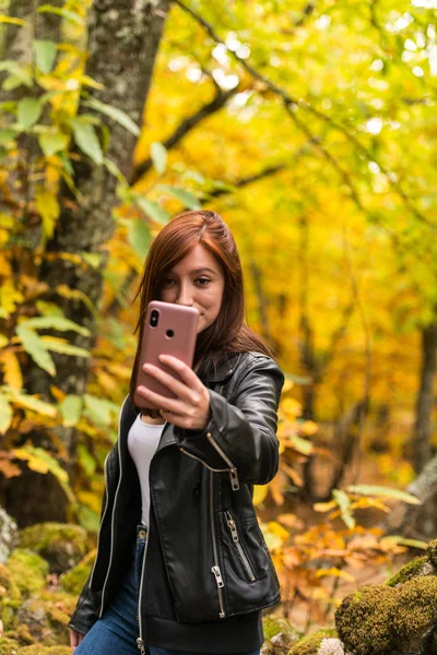 Una joven con el pelo rojizo está haciendo selfies con su teléfono móvil en el bosque — Foto de Stock