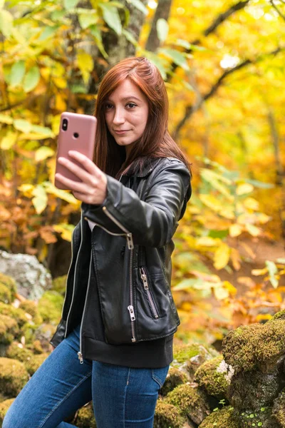Una joven con el pelo rojizo está haciendo selfies con su teléfono móvil en el bosque — Foto de Stock