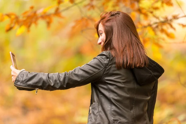 赤味がかった髪を持つ若い女性は森の中の彼女の携帯電話で selfies をやっています。 — ストック写真