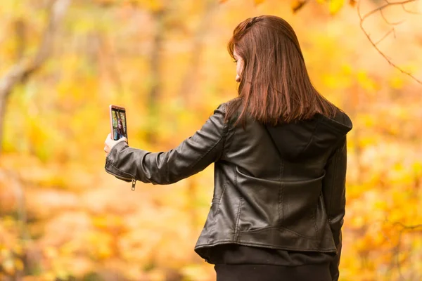 赤味がかった髪を持つ若い女性は森の中の彼女の携帯電話で selfies をやっています。 — ストック写真