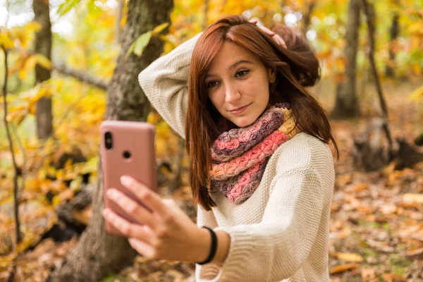 赤味がかった髪を持つ若い女性は森の中の彼女の携帯電話で selfies をやっています。 — ストック写真