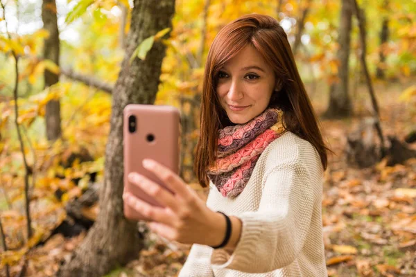 Una joven con el pelo rojizo está haciendo selfies con su teléfono móvil en el bosque — Foto de Stock