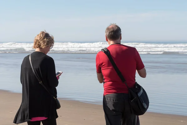 Egy pár turista képeket a telefon és a táblázatok a szénakazalban Rock Cannon Beach, Oregon, Amerikai Egyesült Államok. — Stock Fotó