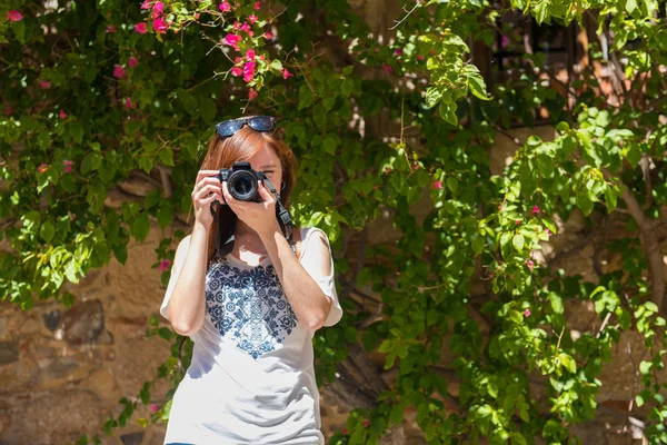 赤みを帯びた髪の若い女性は、ブーゲンビリアカセレスで覆われた壁の隣に彼女のカメラで写真を撮ります — ストック写真