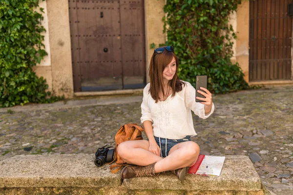 バックパック、カメラ、市の地図を持つ若い赤毛の観光客は、カセレスの旧市街で自分撮りを取ります — ストック写真
