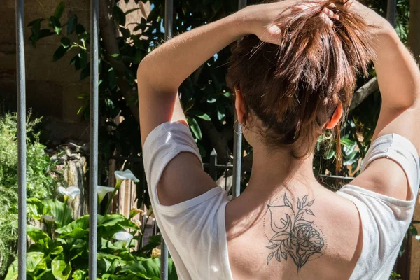 Una joven turista pelirroja con un tatuaje de Arum-lily en la espalda mira un jardín con las mismas flores en Cáceres —  Fotos de Stock