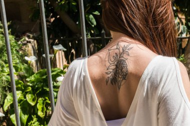 A young red-haired tourist with an Arum-lily tattoo on her back looks at a garden with the same flowers in Caceres clipart