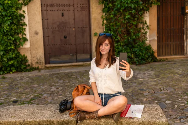 Egy fiatal vörös hajú turisztikai hátizsák, kamera és Várostérkép vesz selfie a régi város csikó — Stock Fotó