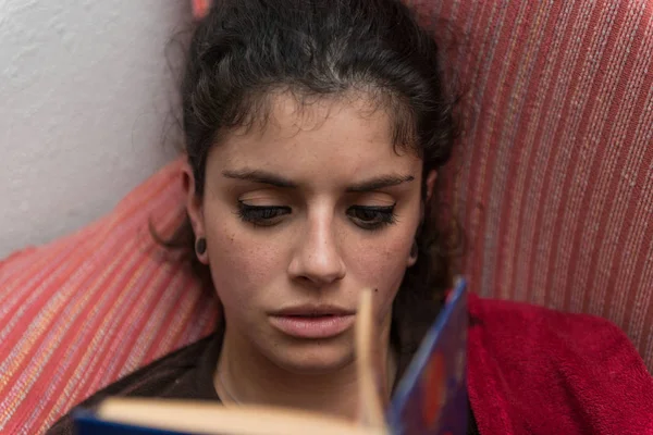 Молодая женщина с каштановыми волосами тихо читает книгу, лежащую на диване, завернутую в красное одеяло . — стоковое фото