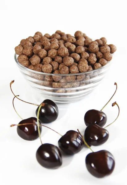 Bolas dulces de chocolate de maíz en un plato de vidrio y bayas de cereza sobre un fondo blanco — Foto de Stock