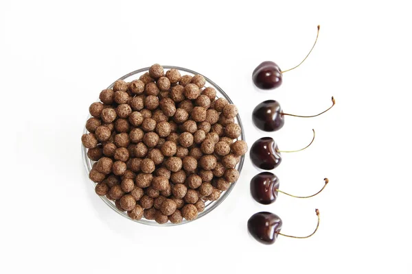 Bolas dulces de chocolate de maíz en un plato de vidrio y bayas de cereza sobre un fondo blanco — Foto de Stock