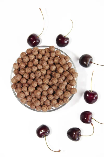 Bolas dulces de chocolate de maíz en un plato de vidrio y bayas de cereza sobre un fondo blanco —  Fotos de Stock