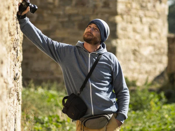 a bearded man wearing a knitted hat stands against a wall in a fortress with a camera in his hand and a camera bag over his shoulder