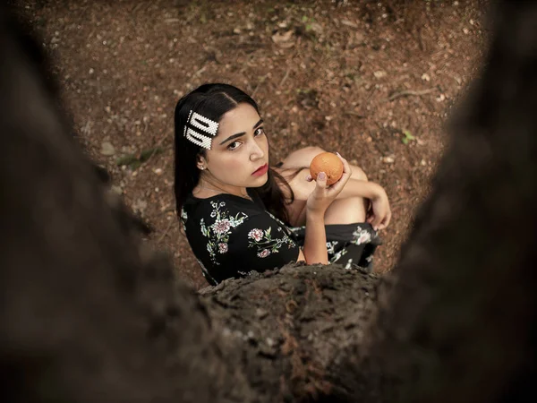 Attractive girl of Caucasian appearance sitting near a tree, holding a tangerine in hand. view from above