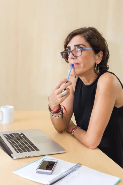 Pensive kvinna med penna i munnen sitter framför sin laptop Stockbild
