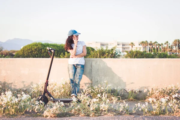 Kadın, elektrikli tekme scooter'ının yanında, cep telefonuyla konuşuyor.