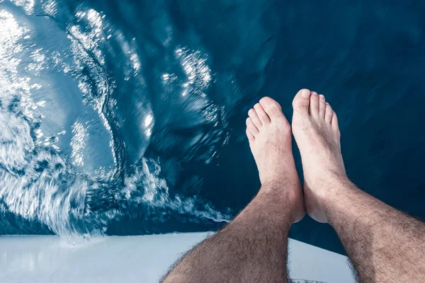 Men\'s feet hanging barefoot over the side of a sailboat. The sailboat\'s on the move.