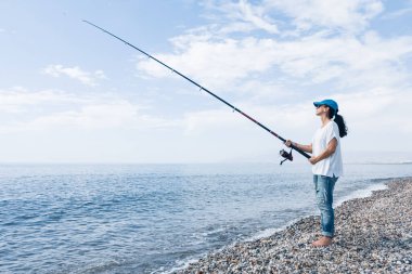 Sahilde balık tutan bir kadın. Güneş gözlüğü ve şapka takıyor..